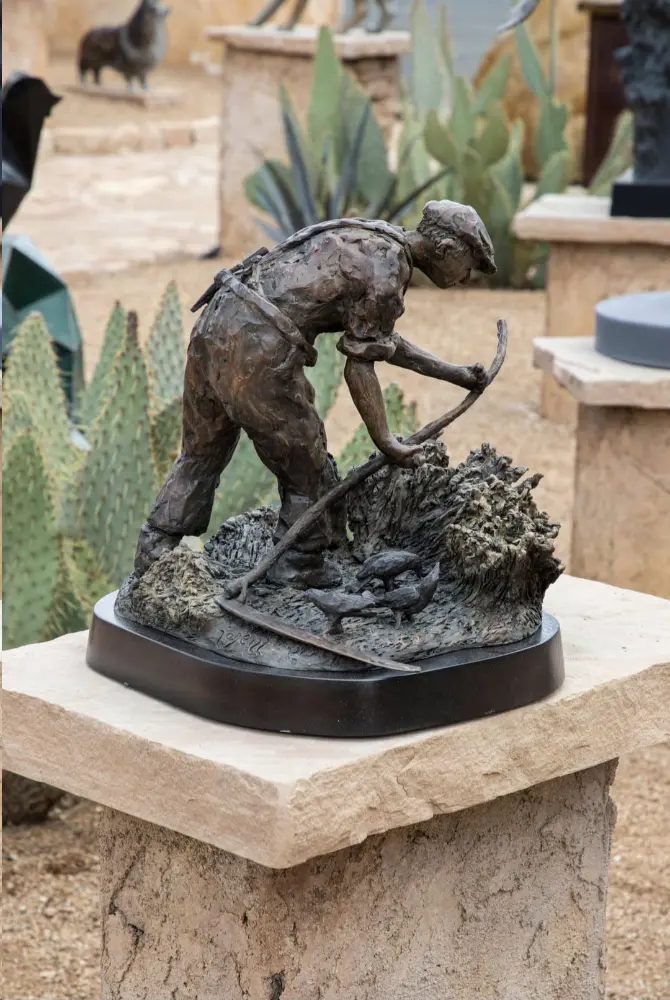 Aged bronze sculpture of a man with a pickaxe working outdoors, set against desert plants and natural stone artworks.